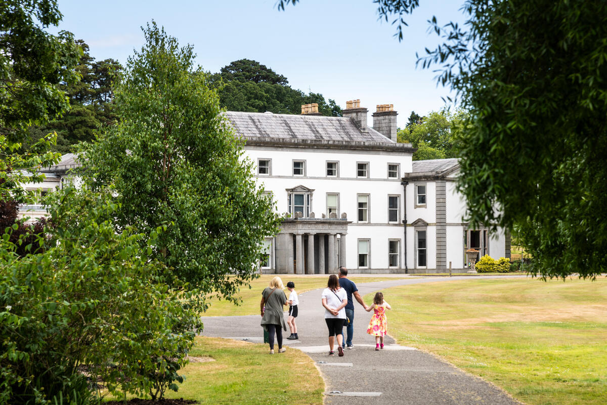 Visitors walking through the grounds towards Fota House in County Cork, with trees framing the historic white house and lawns.